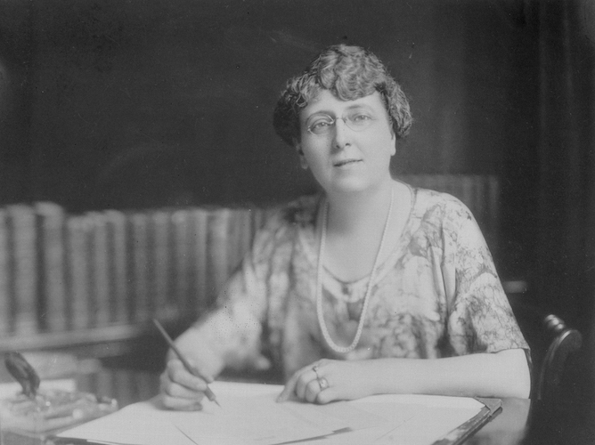Black and white photo of a woman in 1930s clothing sitting at a desk and looking up from a manuscript, a pen in her right hand.
