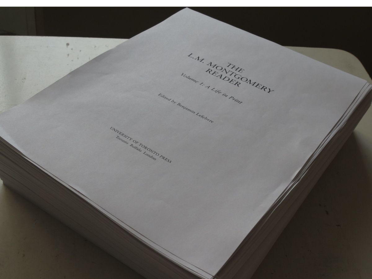 Photo of a stack of pages at the edge of a table. The top sheet reads: "The L.M. Montgomery Reader / Volume 1: A Life in Print / Edited by Benjamin Lefebvre / University of Toronto Press / Toronto, Buffalo, London."