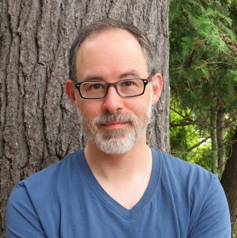 Photo of Benjamin Lefebvre, a Caucasian man with glasses and a salt-and-pepper beard, wearing a blue T-shirt as he stands outside in front of a tree.