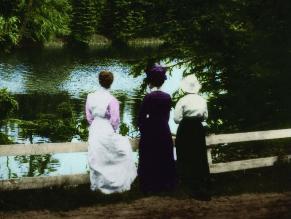 Colourized photograph of three women standing on a dirt road behind a white fence, watching trees on the other side of a body of water.