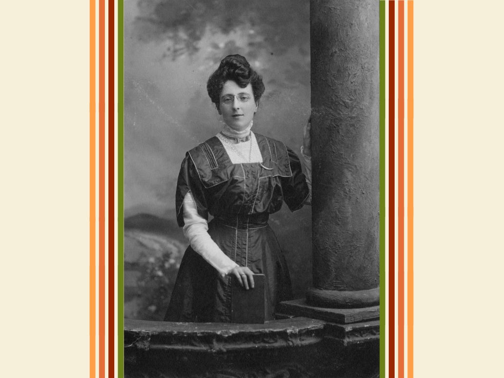 Black and white photograph of L.M. Montgomery, standing, with her left hand touching a column next to her and her right hand resting on a hardcover book on a table in front of her.