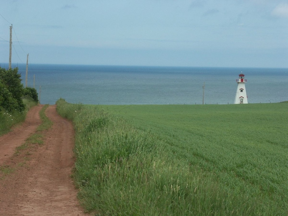 Photo of a PEI landscape, with a red road moving vertically to the left of a green field, with a white lighthouse and the blue ocean in the top third.